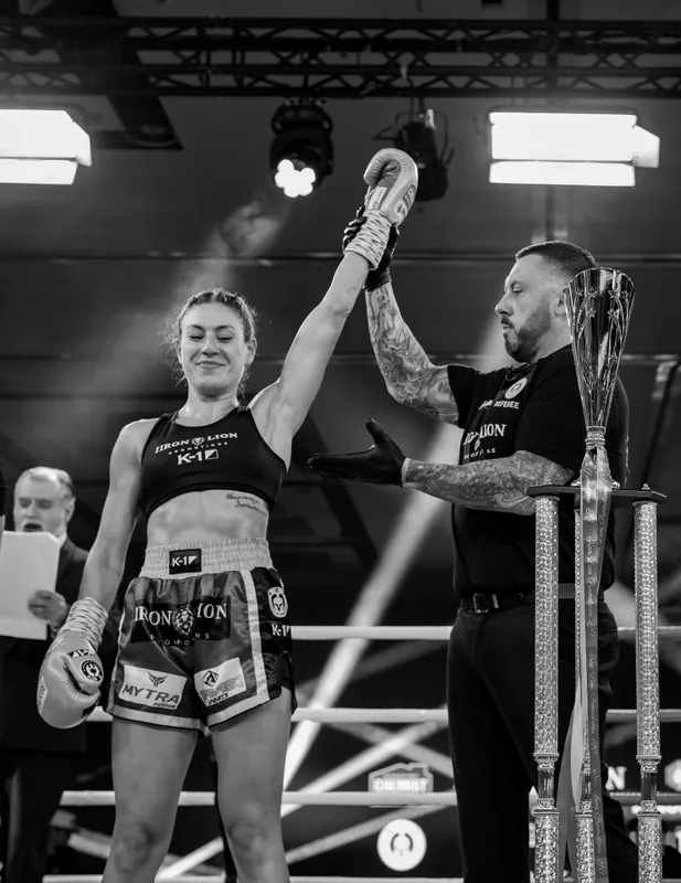 Black-and-white photo of a female Muay Thai fighter having her arm raised by the referee after winning, a trophy beside her