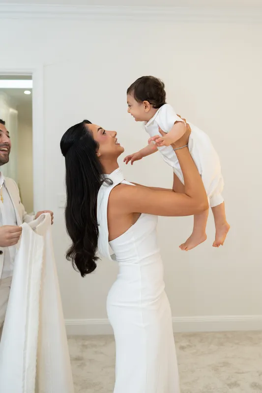 Mother in a white dress lifting her baby boy into the air at his christening