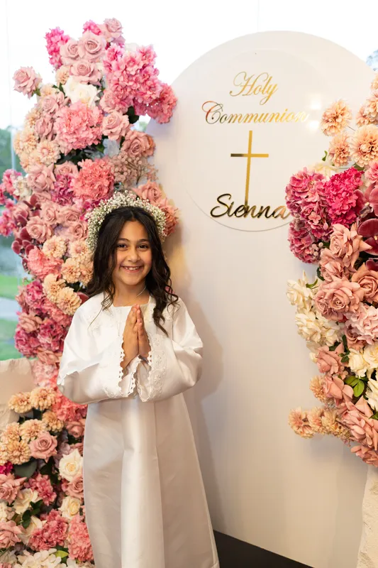 Girl in a white Holy Communion gown praying in front of an arch of pink roses