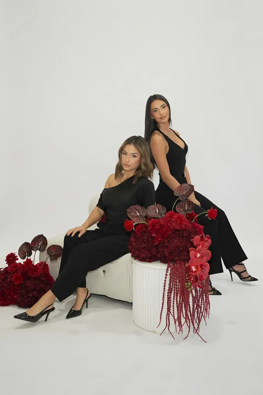 Two Divino Designs founders in black seated beside an arrangement of deep red flowers