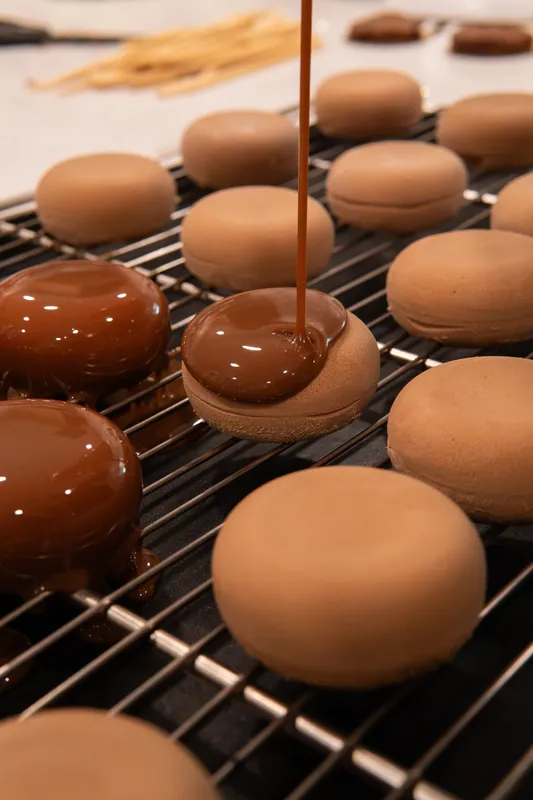 Chocolate glaze being poured over rows of mousse cakes on a cooling rack at Galerie Pâtisserie