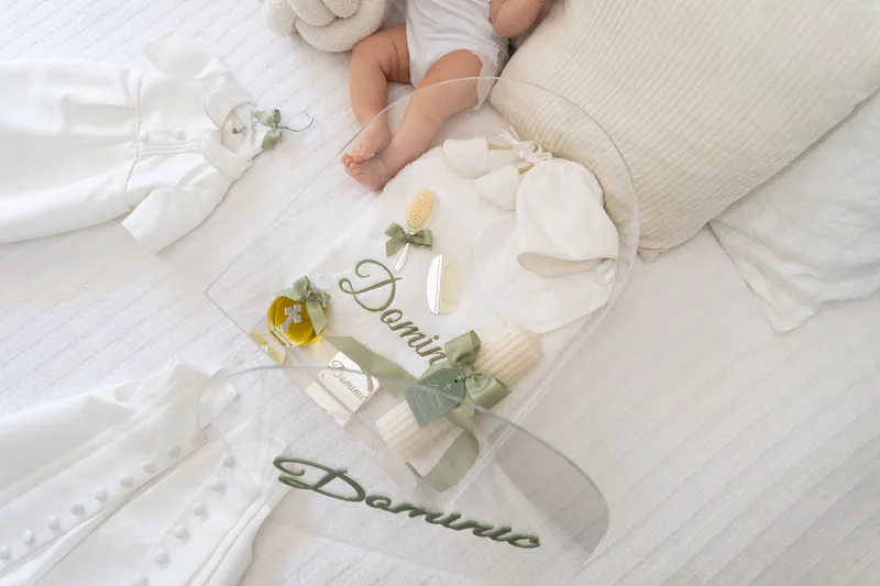 Christening keepsakes laid on white linen beside a sleeping baby: a candle, a cross and an acrylic name plaque