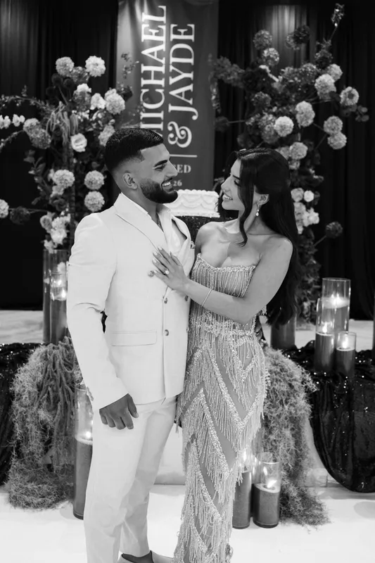 Black-and-white photo of an engaged couple embracing in front of a floral backdrop with candles