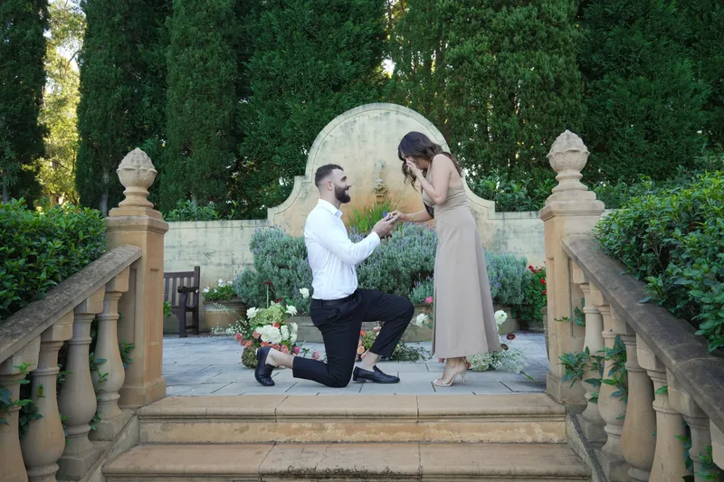 Man on one knee proposing to his partner in a formal garden with hedges and a stone arch