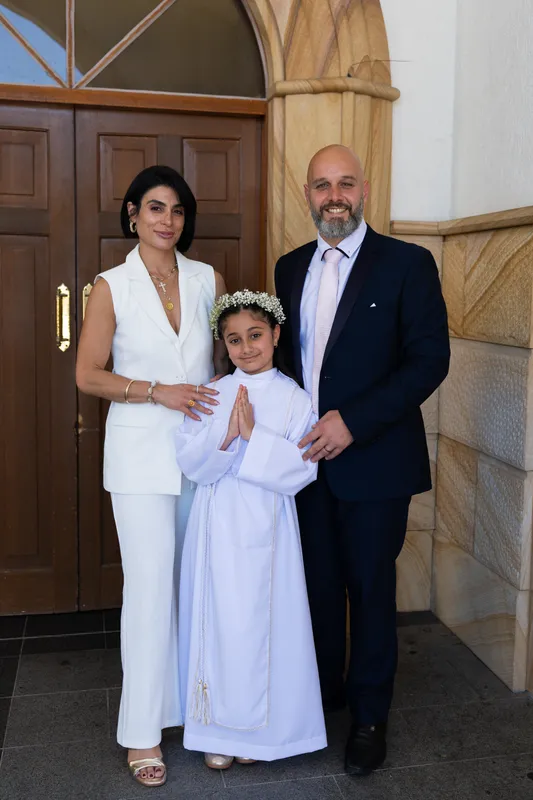 Girl in her white Holy Communion dress and wreath standing with her parents at the church doors