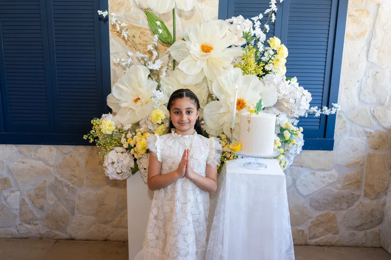 Girl in a white dress with her hands together in front of a floral display at her Holy Communion