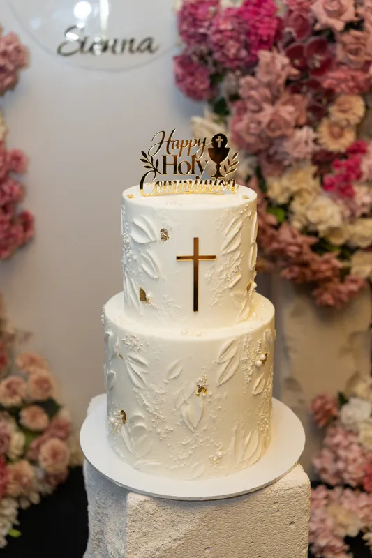 Two-tier white Holy Communion cake with a gold cross in front of pink flowers