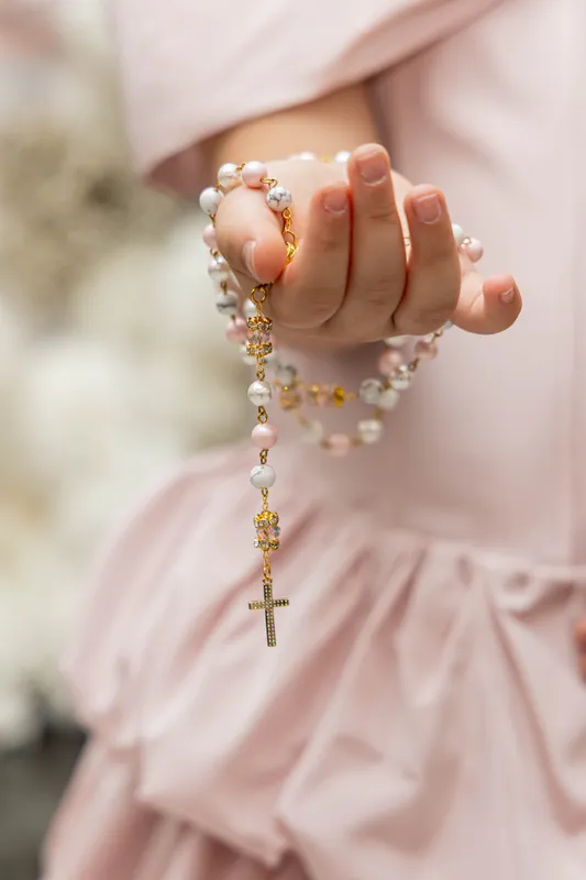 Close-up of a child’s hand holding a pearl rosary against a pink dress