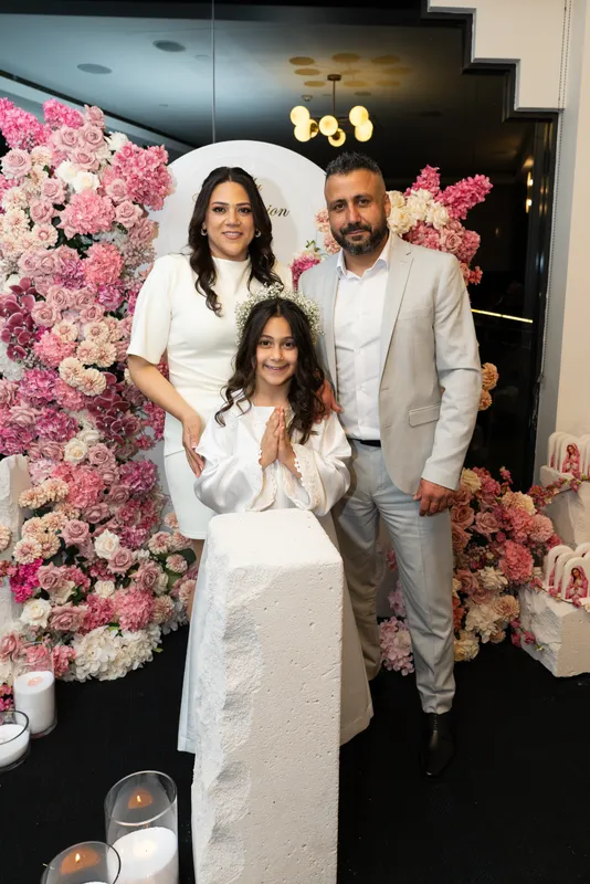 Family photo at a Holy Communion: parents with their daughter in front of a pink floral arch