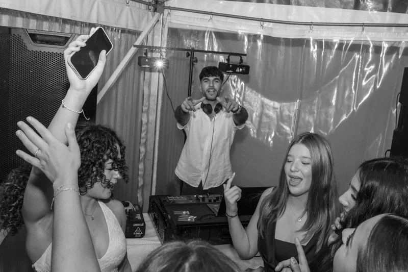 Black-and-white photo of party guests dancing with phones raised while a DJ plays