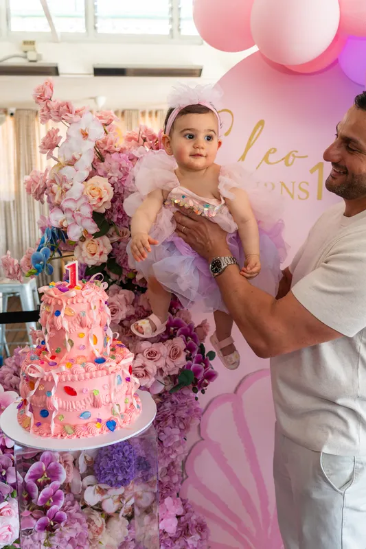 Father holding his baby daughter beside her pink first-birthday cake and flower wall