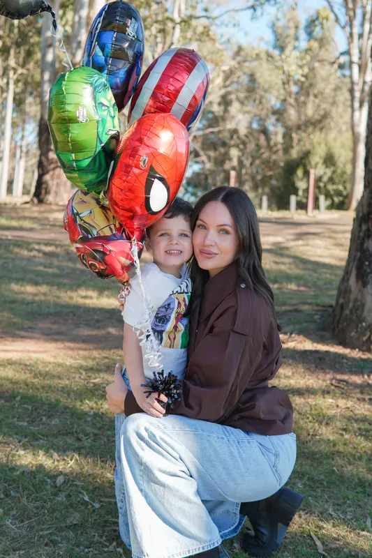 Mother crouching to hug her son beside superhero balloons at an outdoor birthday