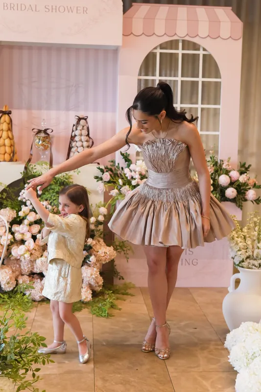 Bride-to-be in a metallic dress at her bridal shower with a little girl beside a pink shopfront backdrop