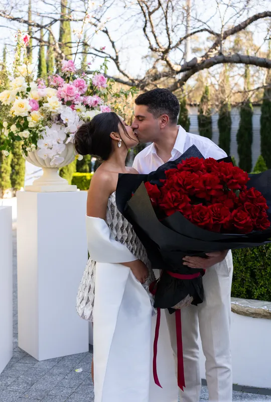 Couple kissing while she holds a large bouquet of red roses at their pre-wedding celebration