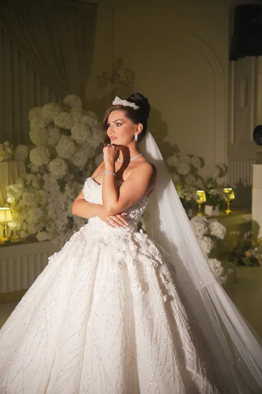 Bride in a beaded gown and long veil posing beside white flowers