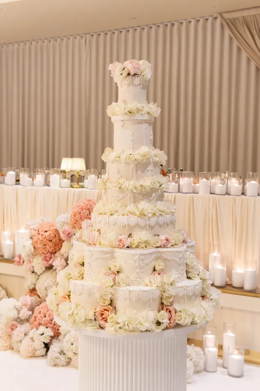 Tall tiered white wedding cake surrounded by peach and white flowers and candles